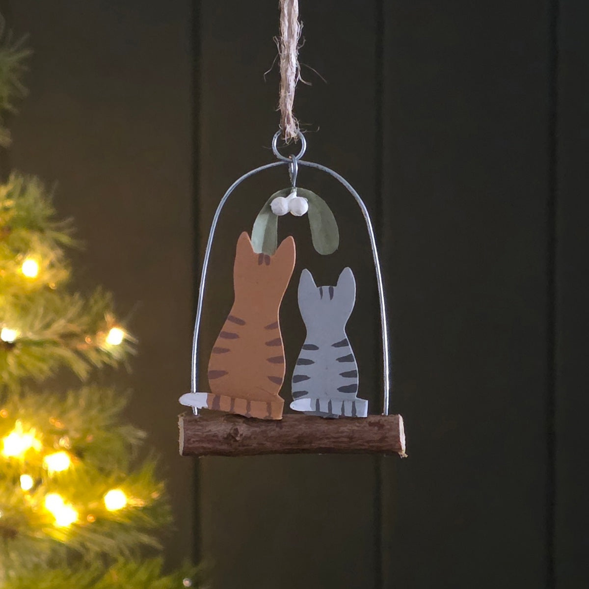 A hanging Christmas ornament shaped like two cats under a wire dome with mistletoe, suspended from a small wooden branch. The ornament is shown against a dark panelled background with blurred warm lights on the left.