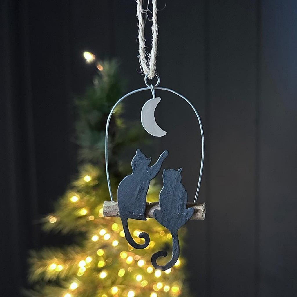 A hanging metal ornament shaped like two black cat silhouettes sitting on a small branch under a silver crescent moon, photographed in front of a blurred Christmas tree with warm lights.