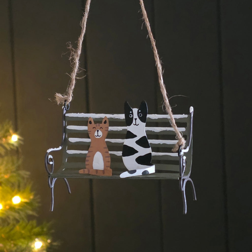 A hanging metal swing bench ornament with two cats painted on it, suspended by twine against a dark panelled background with soft holiday lights on the left.