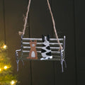 A hanging metal swing bench ornament with two cats painted on it, suspended by twine against a dark panelled background with soft holiday lights on the left.