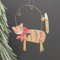 A wooden cat-shaped Christmas ornament with a red and white striped scarf, green holly leaves, and a small gold bell, hanging against a dark grey background with blurred green pine needles on the left.