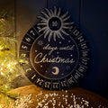 A round wooden navy Christmas advent calendar plaque with a sun, crescent moon, and gold numbered days until Christmas, styled on a wooden surface next to a lit Christmas tree.