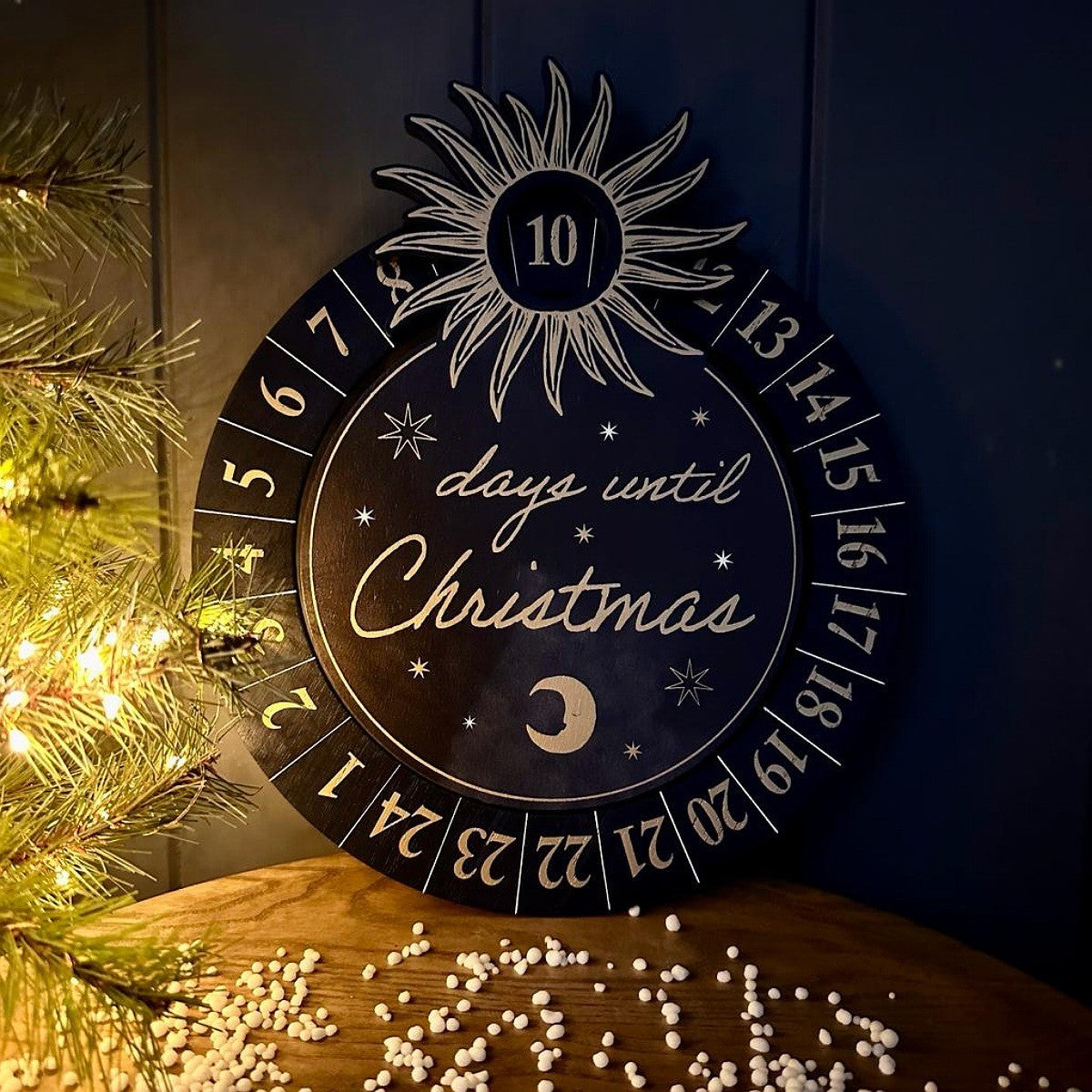 A round wooden navy Christmas advent calendar plaque with a sun, crescent moon, and gold numbered days until Christmas, styled on a wooden surface next to a lit Christmas tree.
