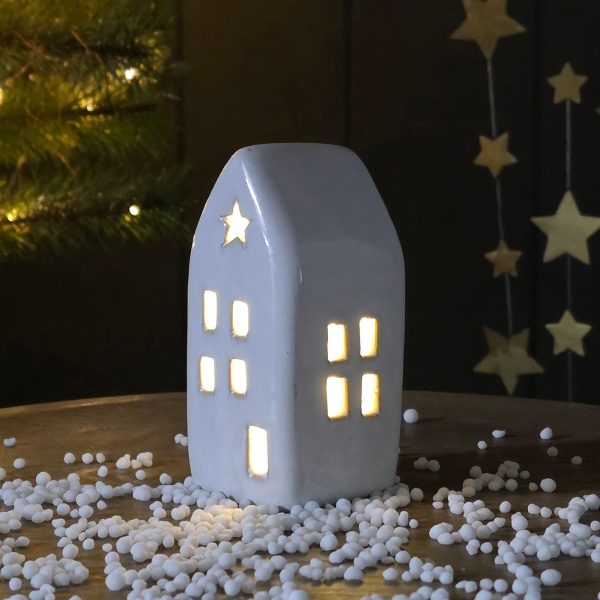 Ceramic house-shaped night light with a star on top, surrounded by decorative snowballs and a Christmas tree in the background.