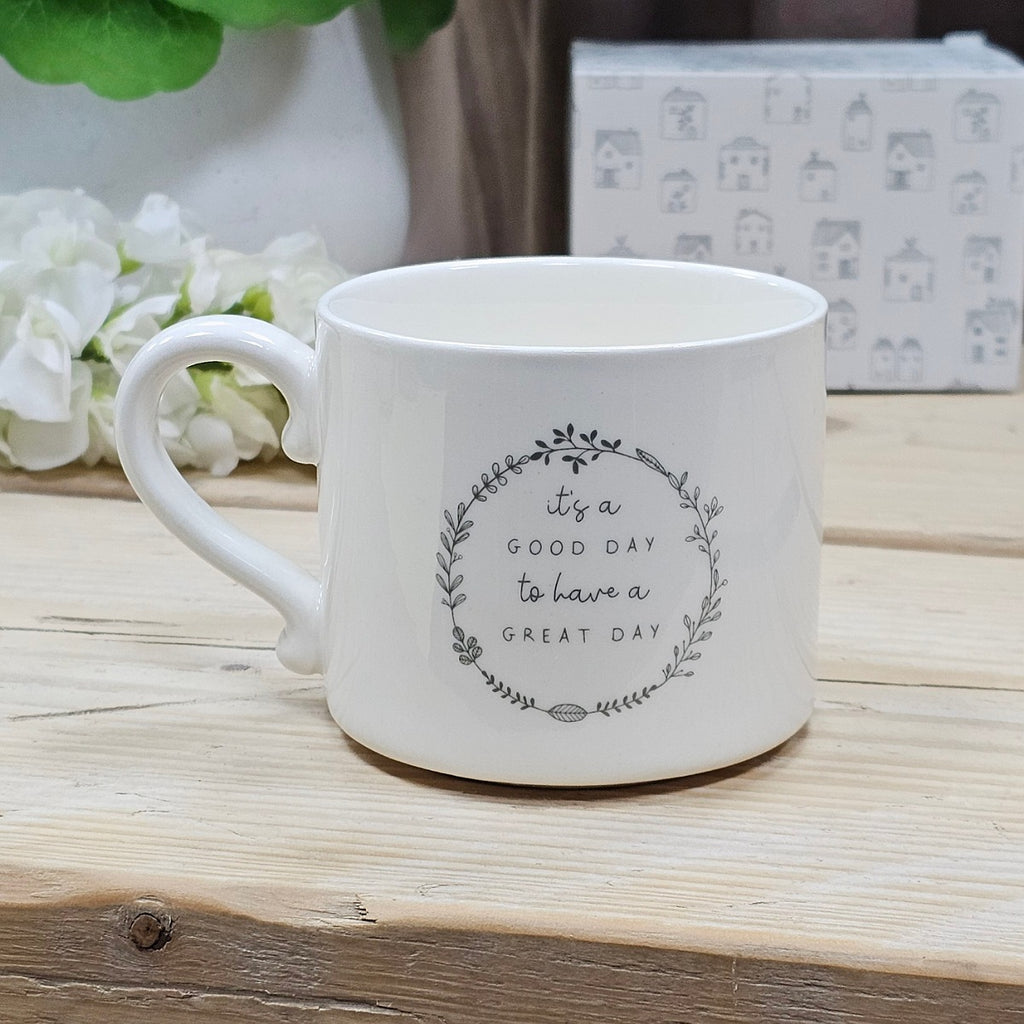 White ceramic mug with the motivational quote 'it's a GOOD DAY to have a GREAT DAY' within a flower wreath on a wooden surface with flowers and a box in the background