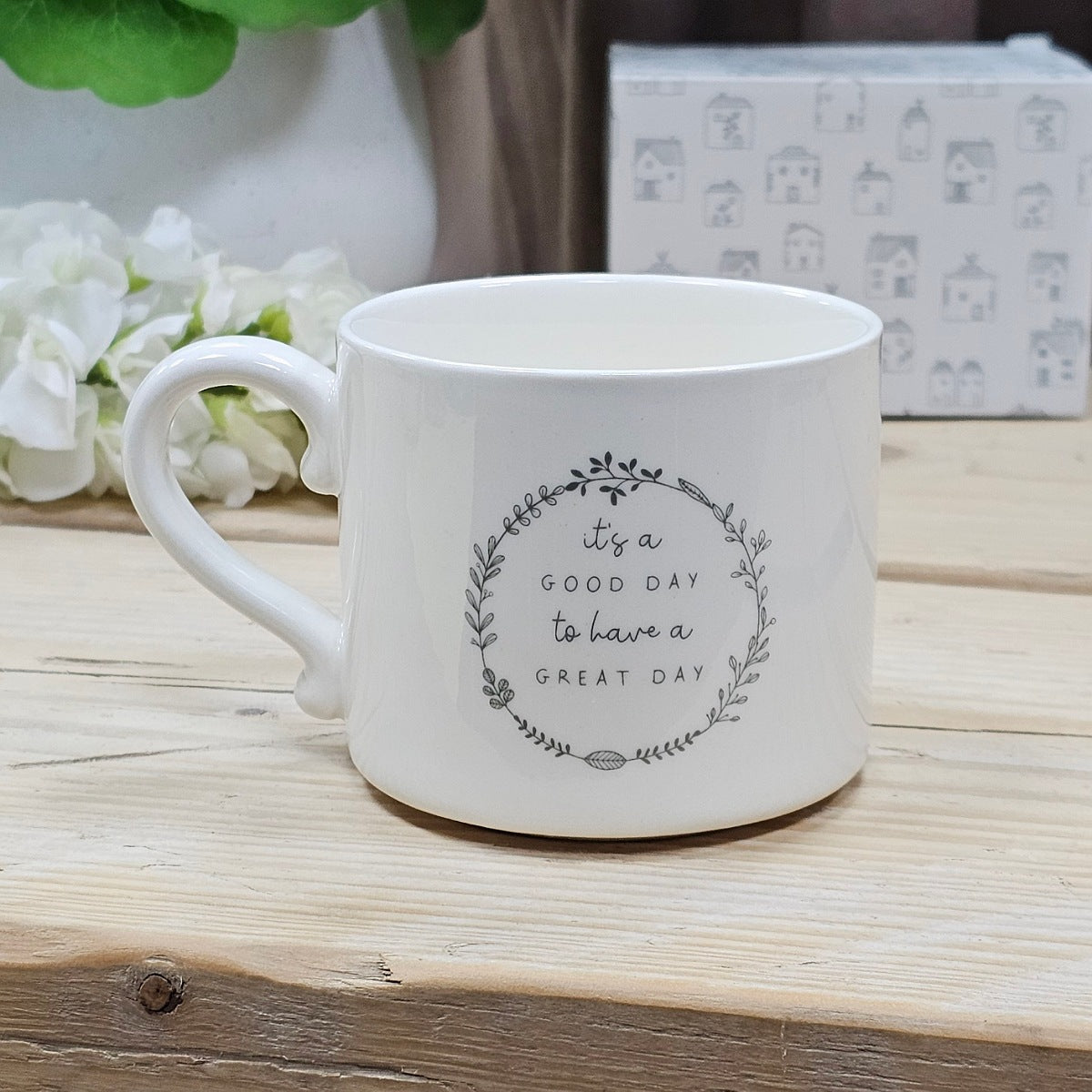 White ceramic mug with the motivational quote 'it's a GOOD DAY to have a GREAT DAY' within a flower wreath on a wooden surface with flowers and a box in the background