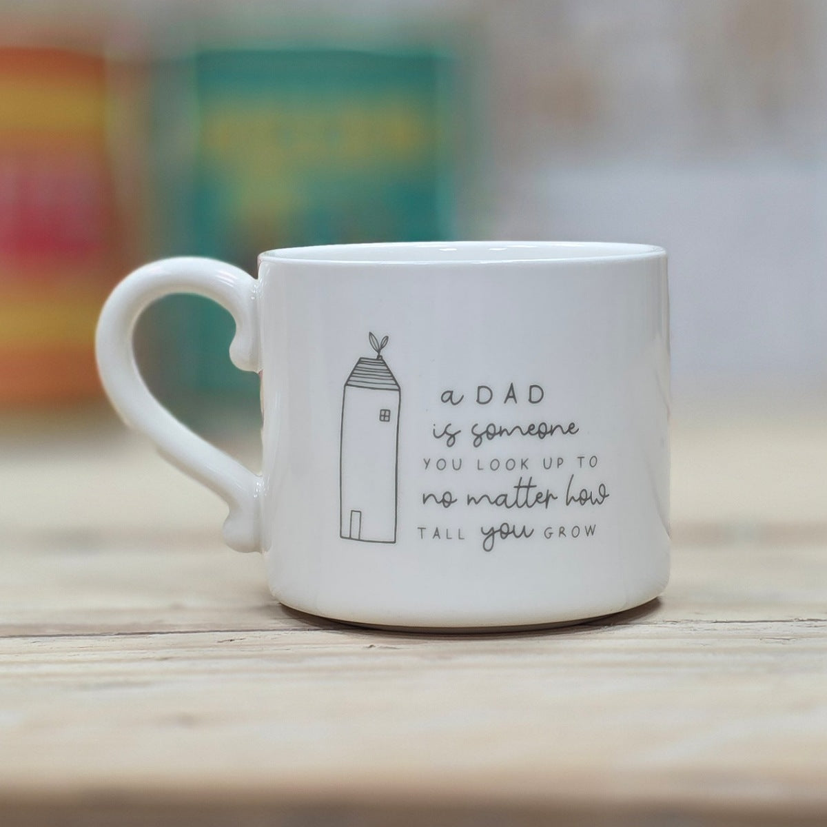 White ceramic mug with the words 'a DAD is someone YOU LOOK UP TO no matter how TALL you grow' with a tall house design to the side, on a wooden surface with a blurred background