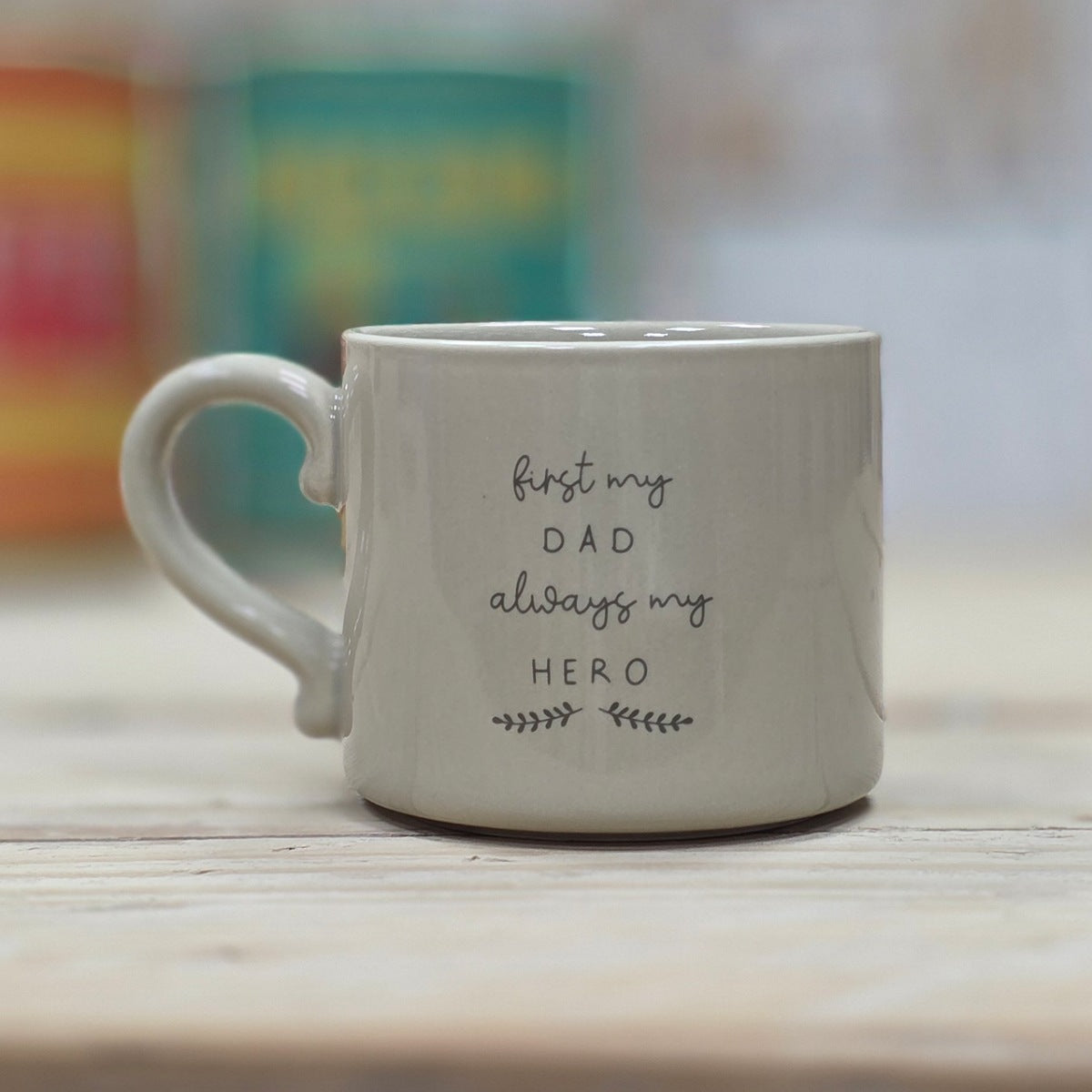 Beige ceramic mug with the words 'first my DAD always my HERO' with a leaf design underneath on a wooden surface and blurred background.