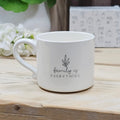 A white ceramic mug with a black printed graphic that reads 'family is EVERYTHING' beneath a small leaf illustration, sitting on a light wooden surface with blurred flowers and a patterned box in the background.