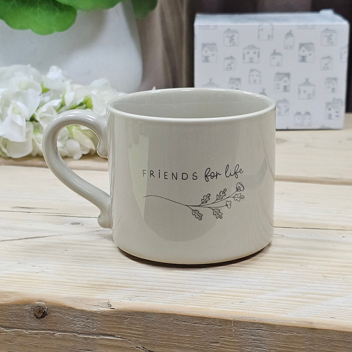 White ceramic mug with the words 'Friends for life' with a branch leaves and flowers underneath on a wooden surface with flowers and a box in the background