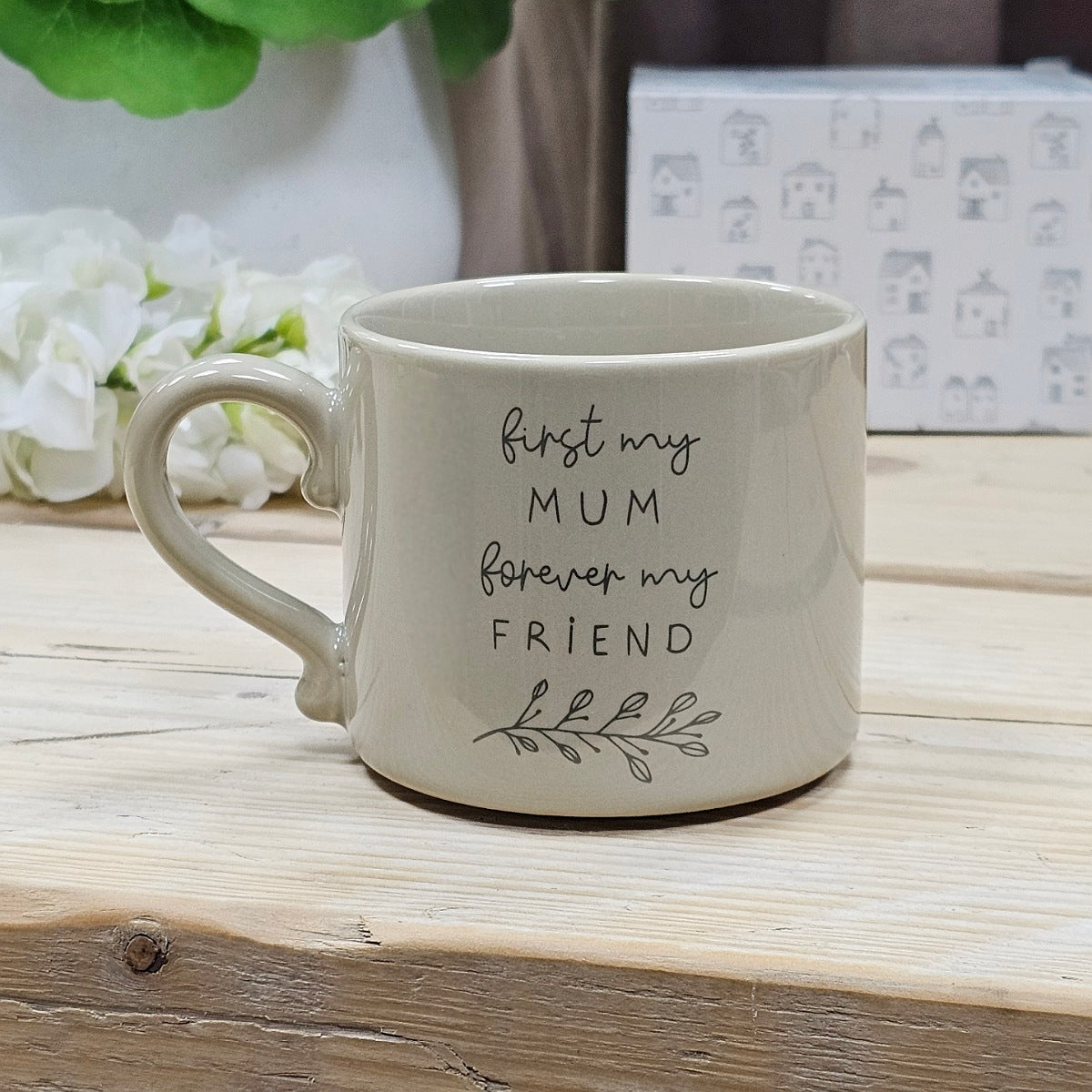 A white ceramic mug with a black printed quote that reads 'first my MUM forever my FRIEND' alongside a simple house illustration, displayed on a wooden surface.