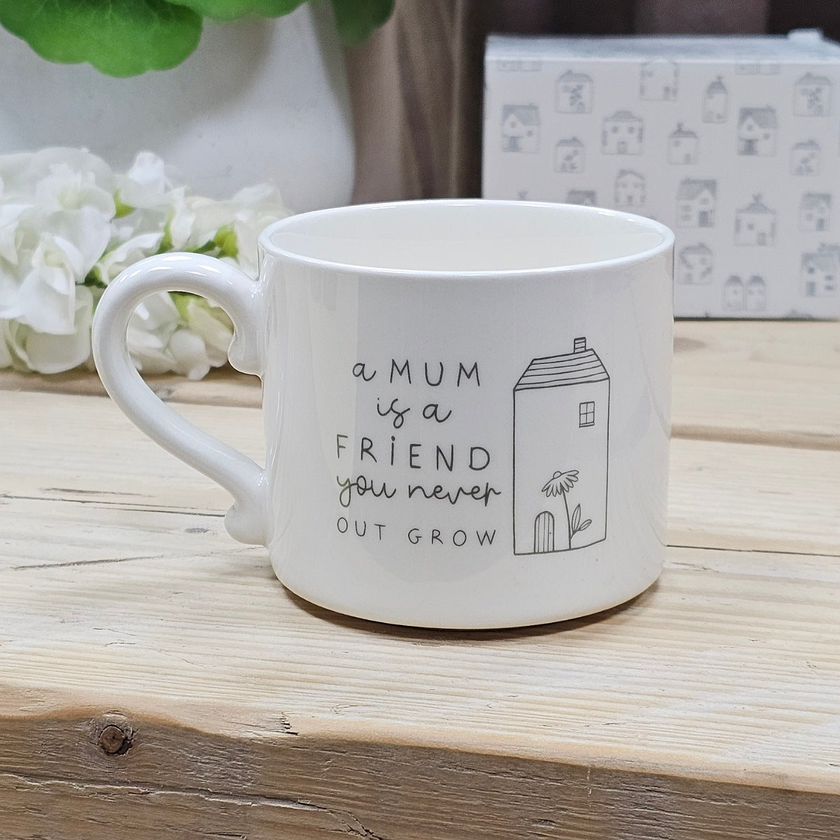 A white ceramic mug with a black printed quote that reads 'a MUM is a FRIEND you never OUT GROW' alongside a simple house illustration, displayed on a wooden surface.