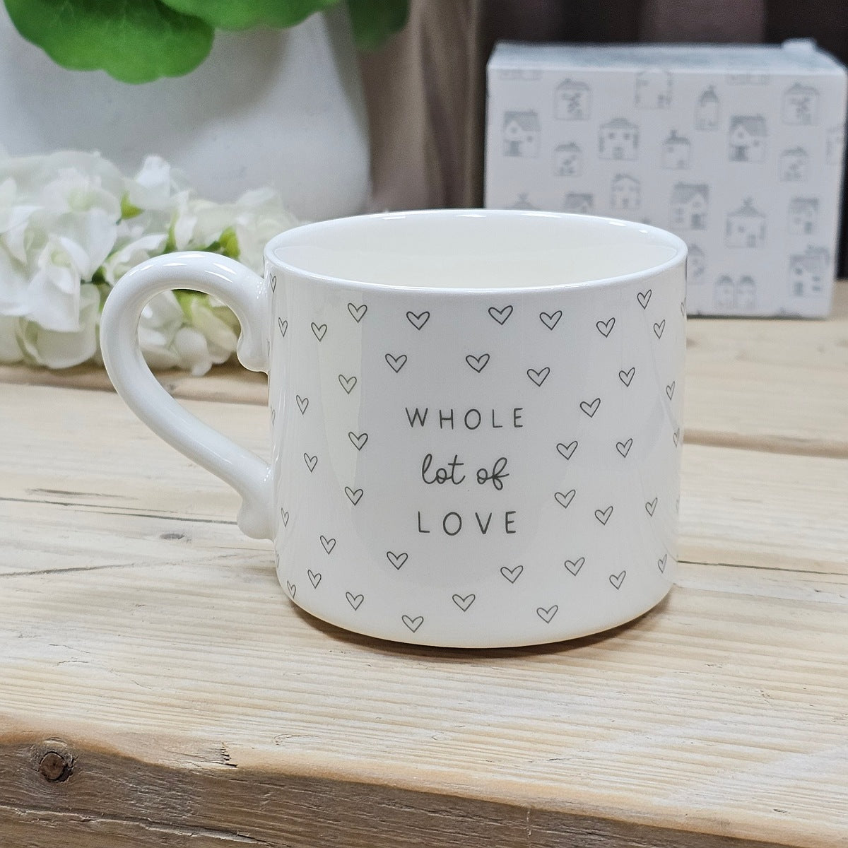 White ceramic mug with the wording 'Whole lot of Love' text and various love hearts on a wooden surface with flowers and a box in the background