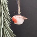 A small hanging ceramic robin ornament with a glossy white body, soft pink-orange face, and brown wings, suspended by a natural twine string against a dark background with blurred greenery on the left.