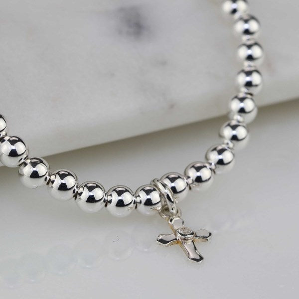 A silver beaded bracelet with a small cross pendant, displayed on a light marble surface.
