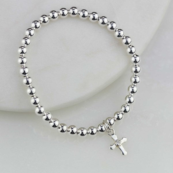 A silver beaded stretch bracelet with a small cross charm, displayed on a white curved surface.
