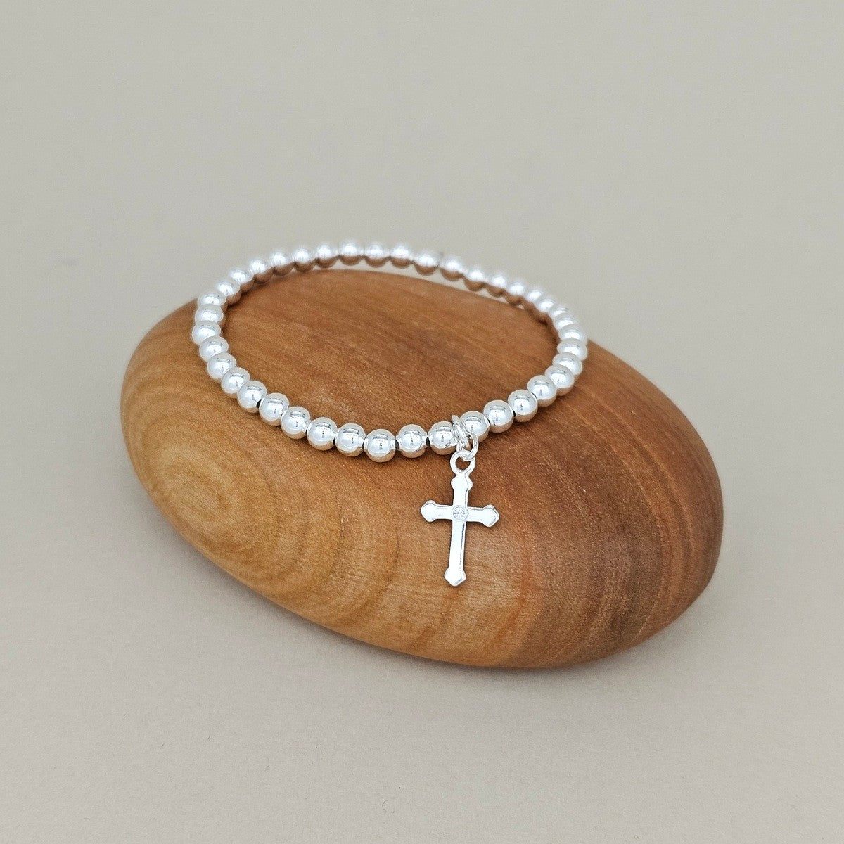 A silver beaded bracelet with a small cross pendant, displayed on a smooth, polished wooden stone against a neutral beige background.