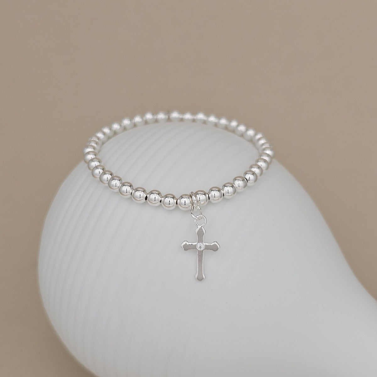 A silver beaded bracelet with a small cross charm, displayed on a white curved stand against a warm beige background.