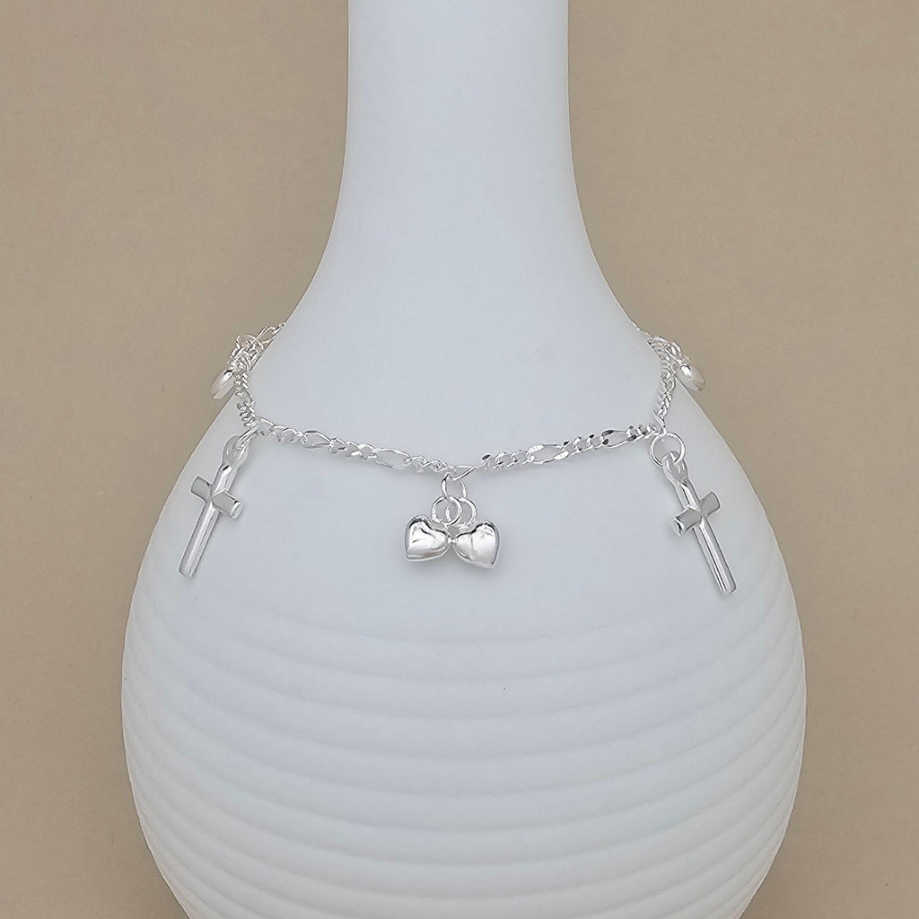 A delicate silver chain bracelet with three small cross charms and a central heart pendant, displayed on a white ribbed ceramic vase against a beige background.