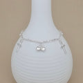 A delicate silver chain bracelet with three small cross charms and a central heart pendant, displayed on a white ribbed ceramic vase against a beige background.