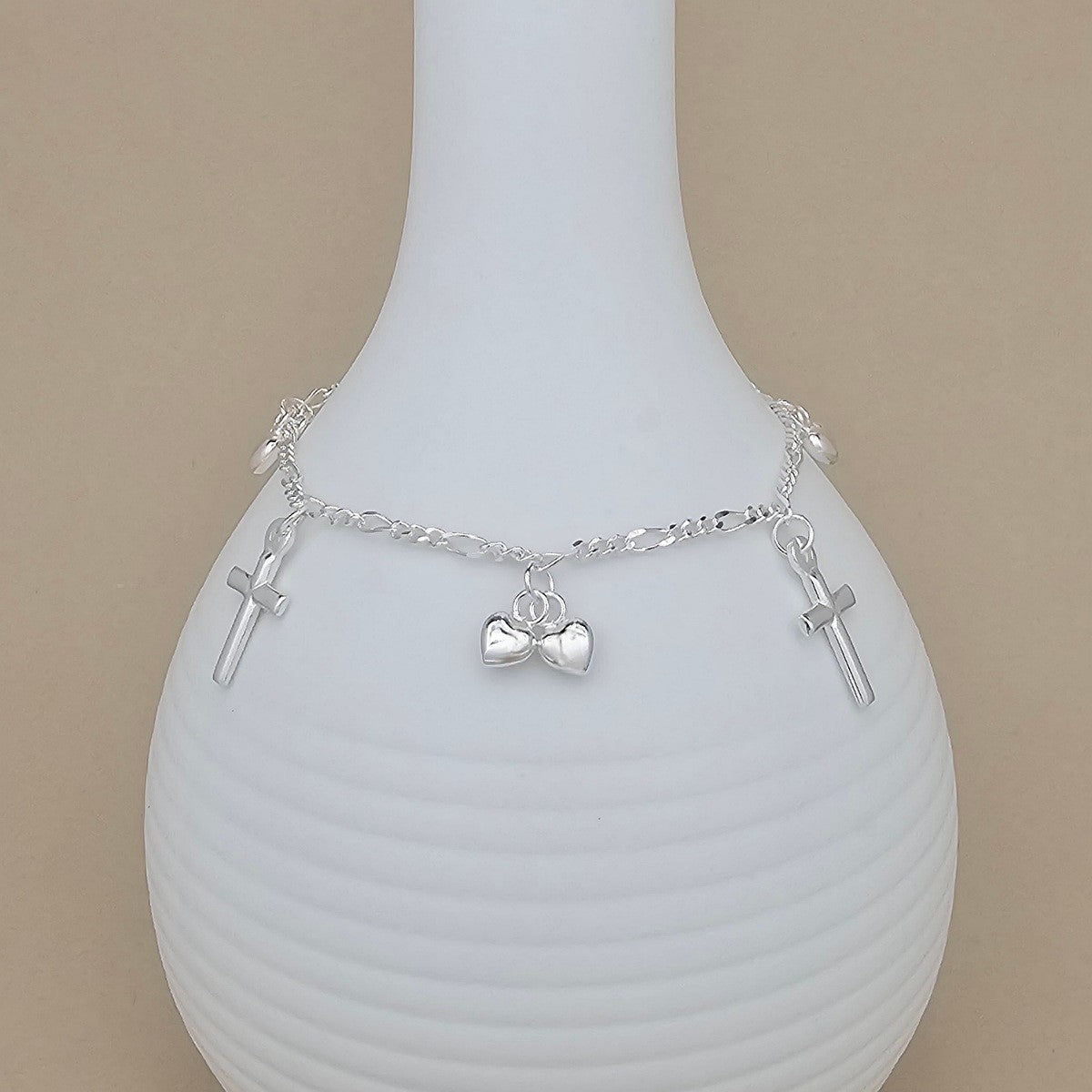 A delicate silver chain bracelet with three small cross charms and a central heart pendant, displayed on a white ribbed ceramic vase against a beige background.