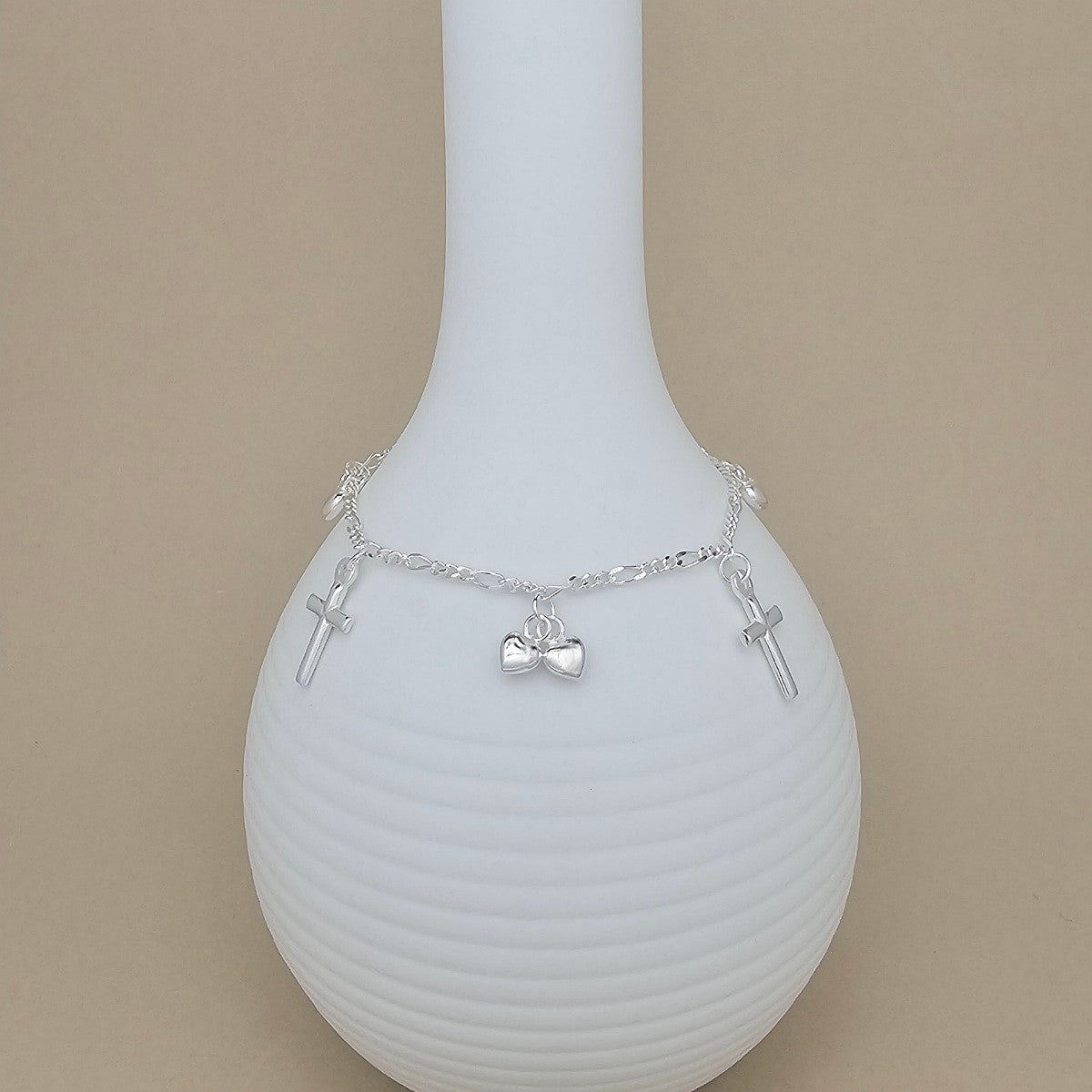 A delicate silver chain bracelet with three charms, including two small crosses and heart charms, displayed on a ribbed white ceramic vase against a solid beige background.