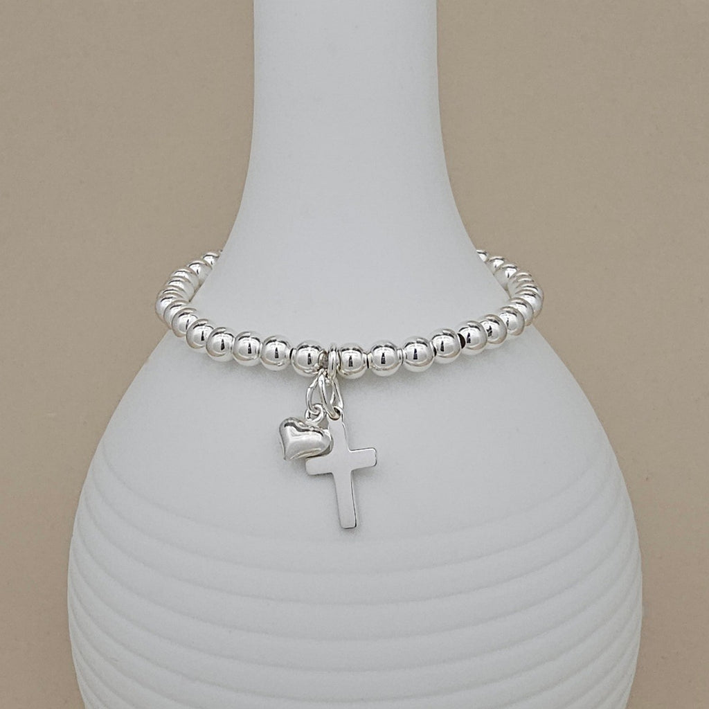 Silver beaded bracelet with a heart and cross charm on a white vase.