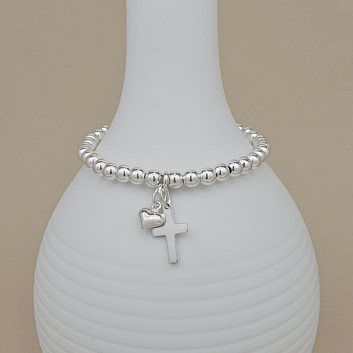 Silver beaded bracelet with a heart and cross charm on a white vase.