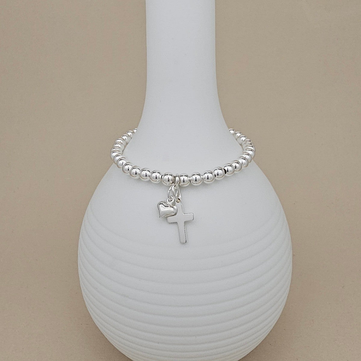 A silver beaded bracelet with a small cross and heart charm, displayed on a white ribbed ceramic vase against a beige background.