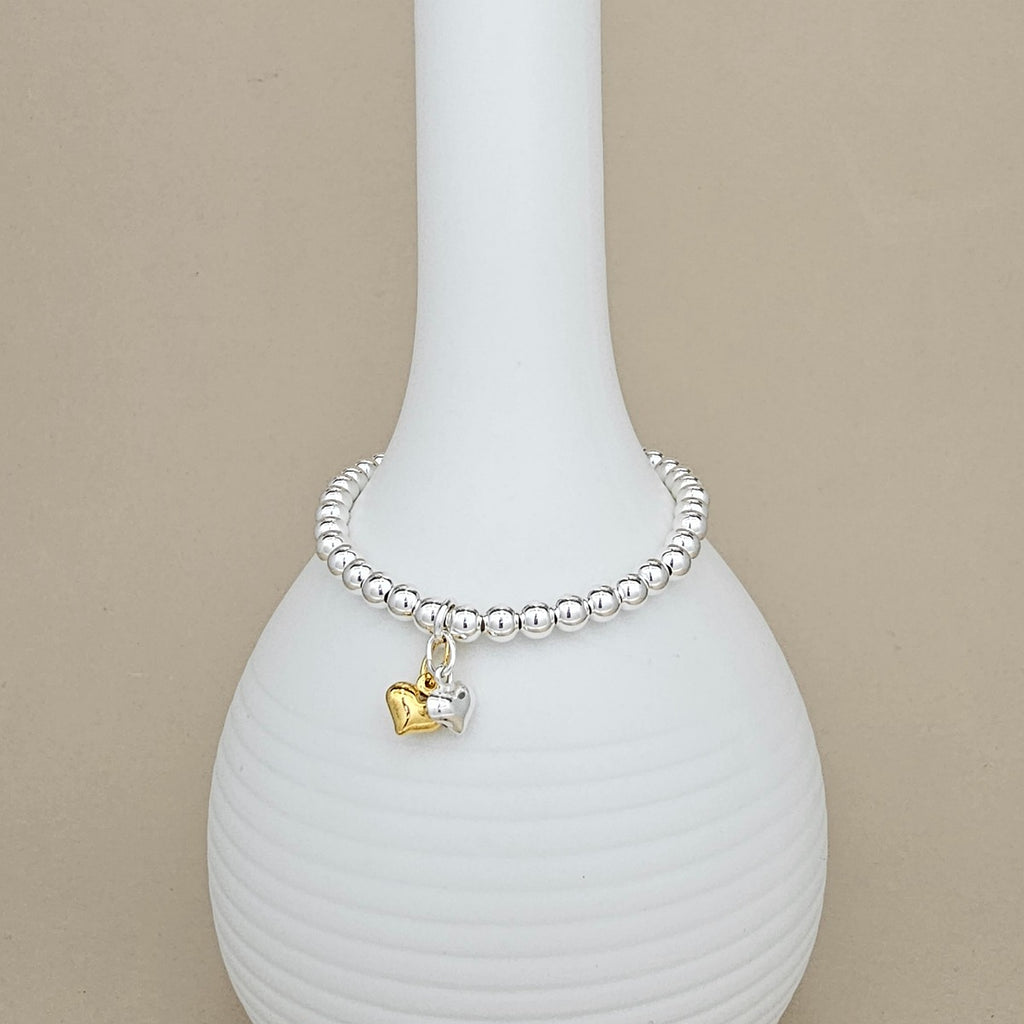 A silver beaded bracelet with a small gold and silver heart charm, displayed on a white ribbed ceramic vase against a beige background.