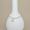 A silver beaded bracelet with a small gold and silver heart charm, displayed on a white ribbed ceramic vase against a beige background.