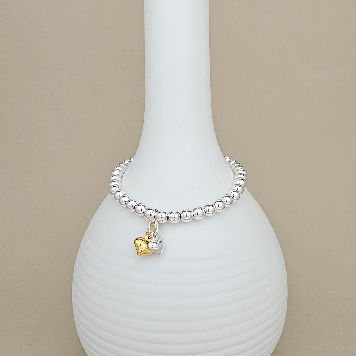 A silver beaded bracelet with a small gold and silver heart charm, displayed on a white ribbed ceramic vase against a beige background.