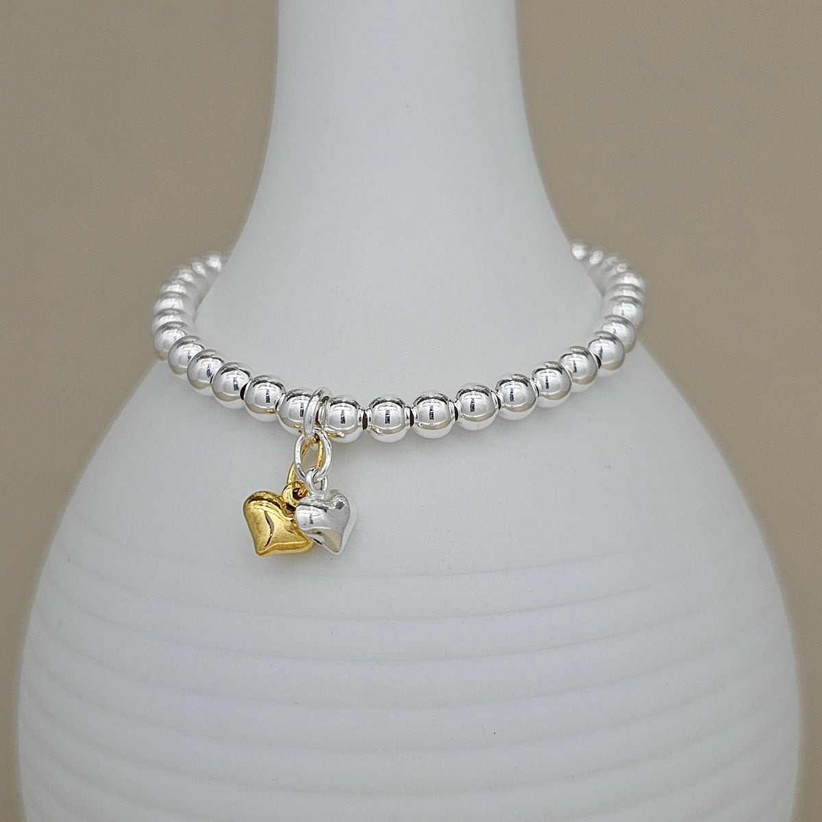 A silver beaded bracelet with two heart charms, one gold and one silver, displayed on a white ceramic vase against a neutral beige background.
