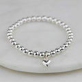 A polished silver beaded bracelet with a small dangling heart charm, displayed on a white marble surface.