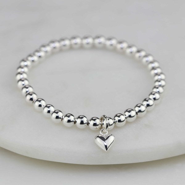 A polished silver beaded bracelet with a small dangling heart charm, displayed on a white marble surface.