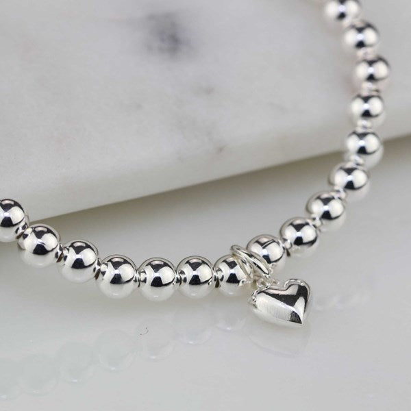 A close-up of a silver beaded bracelet with a small polished heart charm, resting on a white marble surface.