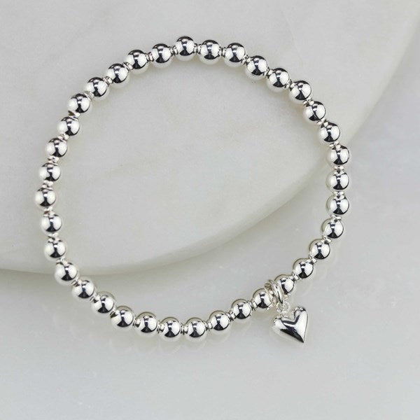 A silver beaded bracelet with a small dangling heart charm, displayed on a white curved surface.