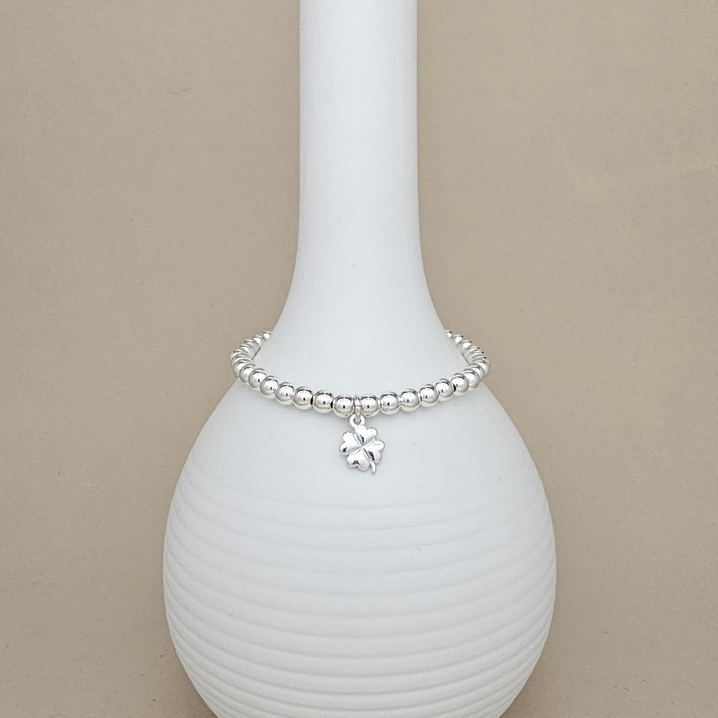A silver beaded bracelet with a small clover charm, displayed on a white ribbed ceramic vase against a beige background.