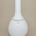 A silver beaded bracelet with a small clover charm, displayed on a white ribbed ceramic vase against a beige background.