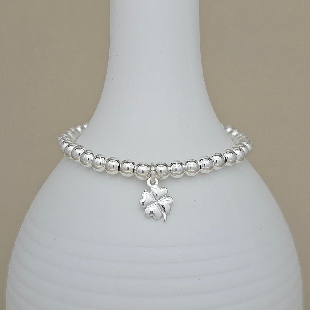 A silver beaded bracelet with a small four-leaf clover charm, displayed on a white ceramic vase against a neutral beige background.