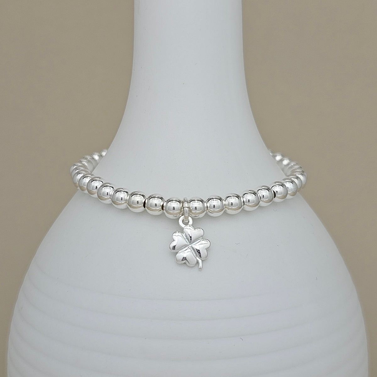 A silver beaded bracelet with a small four-leaf clover charm, displayed on a white ceramic vase against a neutral beige background.
