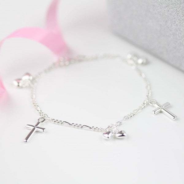 A delicate silver chain bracelet with three dangling cross charms and two small heart charms, displayed on a soft white surface with a pink ribbon and a gray box in the background.