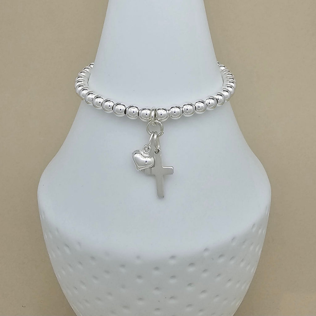 A silver beaded bracelet with a small cross and heart charm, displayed on a white ceramic jewelry bust against a beige background.