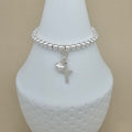 A silver beaded bracelet with a small cross and heart charm, displayed on a white ceramic jewelry bust against a beige background.