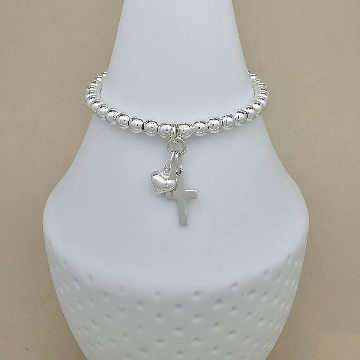 A silver beaded bracelet with a small cross and heart charm, displayed on a white ceramic jewelry bust against a beige background.