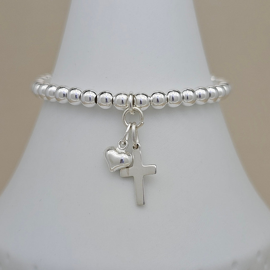 A silver beaded bracelet with a dangling cross and small heart charm, displayed on a white jewelry bust against a soft beige background.