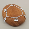 A silver chain bracelet with small charms, including two crosses and small hearts, displayed on a smooth brown wooden stone against a light beige background.