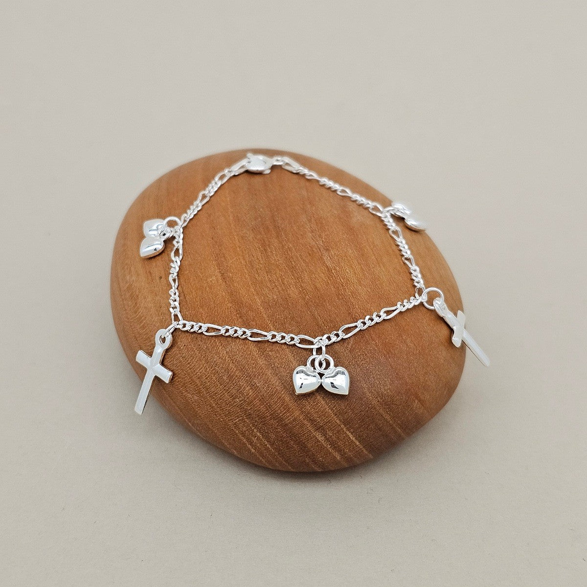 A delicate silver chain bracelet with small charms, including two crosses and heart charms, displayed on a smooth, oval-shaped wooden stone against a neutral beige background.
