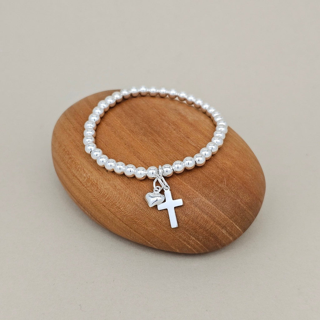A beaded stretch bracelet with silver beads and a silver cross and heart  charm, displayed on a smooth oval wooden block against a neutral beige background.