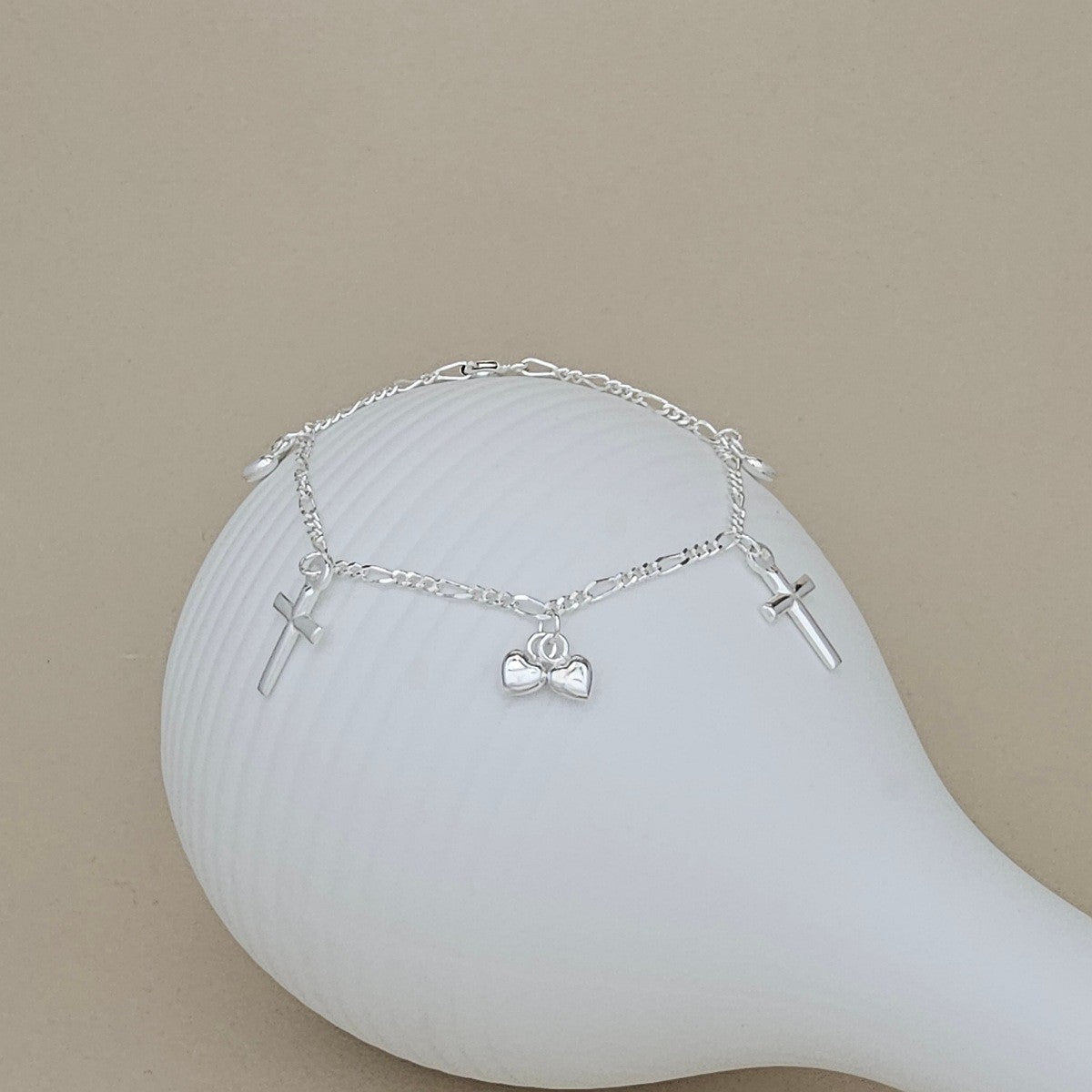 A silver chain bracelet with three charms, including two small crosses and 2 hearts, displayed on a white ceramic mannequin against a beige background.